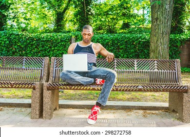 College Student Studying in Summer in New York. Wearing black, white striped tank top, jeans, red sneakers, an African American guy sitting on chair on campus, reading, working on laptop computer.
