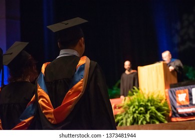 College Students Dressed In Cap And Gowns Listen To A Speech During A Graduation Ceremony.