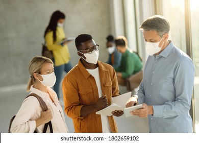 College Students And Their Teacher Wearing Protective Face Masks While Using Touchpad In A Hallway. Focus Is On African American Student. 