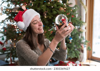 College-aged woman looking at Christmas snow-globe by Christmas Tree - Powered by Shutterstock