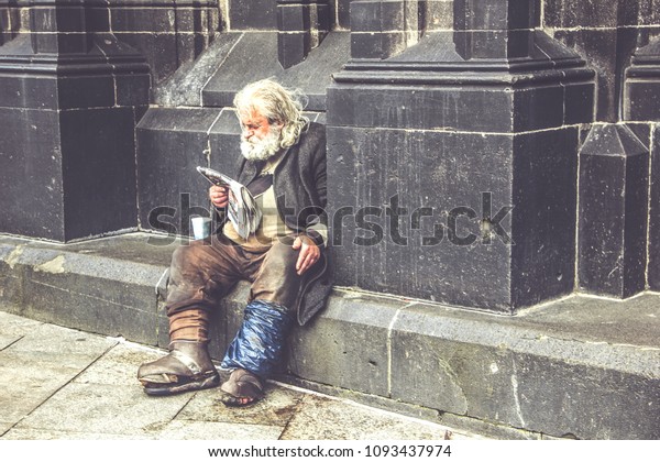 COLOGNE, GERMANY\
- April 16, 2018: A homeless tramp reads a newspaper sitting on the\
street near the\
cathedral