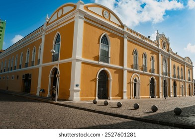 colonial style building with a yellow facade and white trim, arched windows and an ornate white cornice along the roofline, foreground a cobblestone street with spherical balls, background a blue sky - Powered by Shutterstock