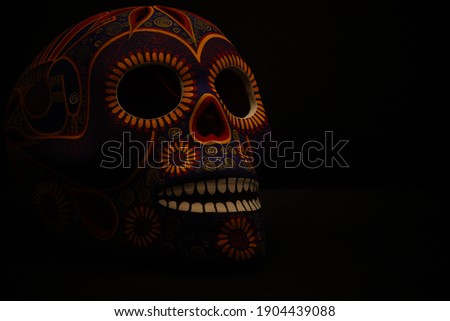 color photograph of a Mexican catrina colored in red, lilac, blue and yellow. The skull is looking to the right. Black background