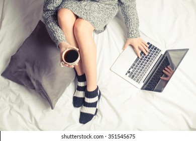 Color picture of a beautiful young woman drinking coffee at home in her bed wearing a cozy sweater while checking her laptop