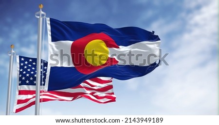 The Colorado state flag waving along with the national flag of the United States of America. In the background there is a clear sky.