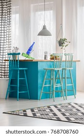 Colored bar stools at blue table in bright dining room with patterned carpet, grey lamp and bottles