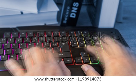 Colored computer keyboard with human hands in motion. Close-up of writing fingers of hacker, spy or censor. Detail of a book with Secure DNS lettering on background. Digitization and cyber security.