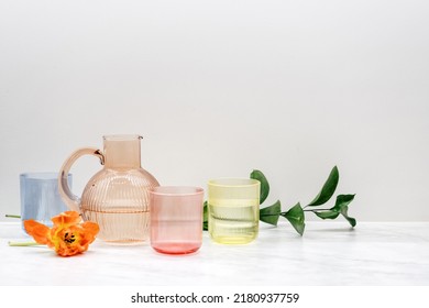 Colored Jug And Transparent Glasses With Water. Stylish Kitchen Tableware And Flowers. Minimalism Style. Home Comfort. Objects Composition Idea