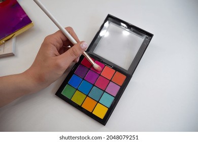 Colored Makeup Palette Lies On The Table. The Brush Held By The Makeup Artist Is Dipped To Pick Up Color