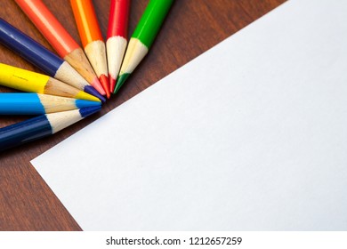 Colored pencils in array on desk with white paper background.