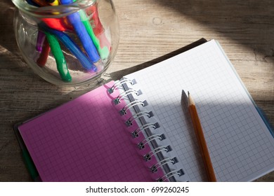 Colored pencils and pens in a glass jar and an open notebook on a wooden table - Powered by Shutterstock