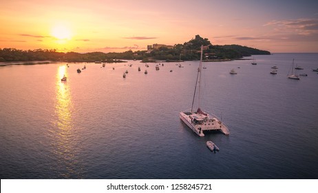 A colorful aerial shot of a large white catamaran yacht anchoring in a calm bay while watching a beautiful sunset on the coast of Costa Rica in Central America.