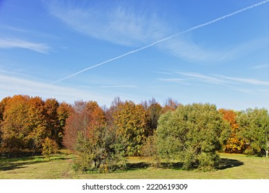 Colorful Autumnal Trees In The European Park At Sunny October Day On Blue Sky With Clouds And Plane Vapor Exhaust Background