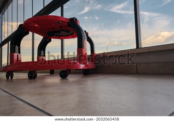 Colorful\
baby-walker on faience floor inside the huge balcony with cloudy\
sky background in glass made fences in\
balcony.