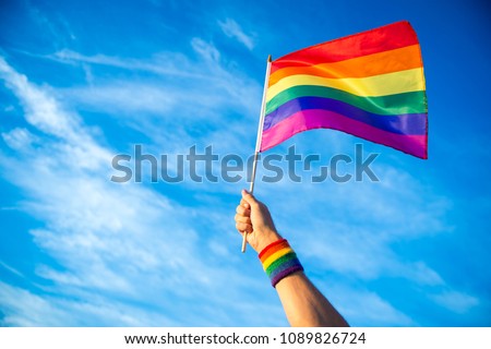 Colorful backlit rainbow gay pride flag being waved in the breeze against a sunset sky.