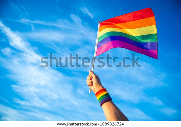 Colorful backlit rainbow gay pride flag being\
waved in the breeze against a sunset\
sky.