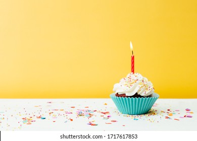 A colorful  birthday cupcake with one candle and confetti on a yellow background - Powered by Shutterstock