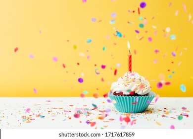 A colorful  birthday cupcake with one candle and confetti on a yellow background