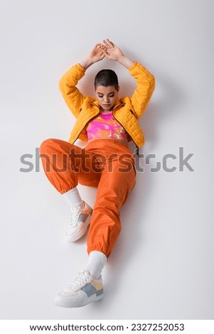 colorful clothes, outerwear, female model posing in casual attire, young woman with short hair and puffer jacket sitting with raised hands on grey background, personal style, self expression