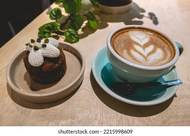 Colorful Cup With Latte Coffee With Flower Design, Delicious Gourmet Cake With Cream And Green Decorative Plant In A Coffee Shop Interior In The Night