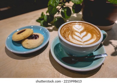 Colorful Cup With Latte Coffee With Flower Design, Delicious Homemade Cookies And Green Decorative Plant In A Coffee Shop Interior In The Night