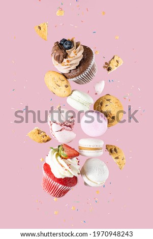 Colorful decorated Broken chocolate chip cookies, cupcakes and macaroons falling in motion on pink background with sprinkling and pieces with crumbs. Sweet and various pastries flying.