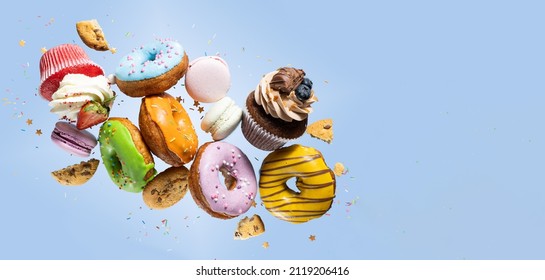 Colorful decorated donuts, cupcakes and macaroons falling in motion on blue background with sprinkling. Sweet and various doughnuts flying on pastel backdrop. Banner - Shutterstock ID 2119206416