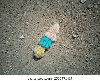 a colorful duster with a striped handle, lying on a dirt and rocky surface. The duster's head is made of soft fibers in different colors: pink, blue, and yellow. - Powered by Shutterstock