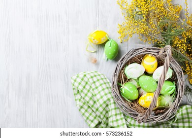 Colorful easter eggs in basket and mimosa flowers on wooden table.  Top view with copy space 