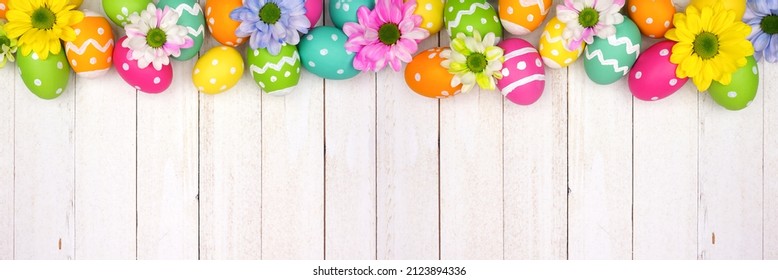Colorful Easter Eggs And Spring Flowers. Overhead View Top Border Against A White Wood Banner Background. Copy Space.