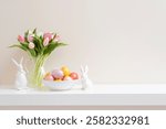 Colorful easter eggs, spring tulip flowers and ceramic bunny figurines on white table over plain wall background