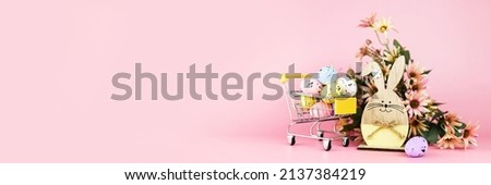 Colorful easter quail eggs in shopping cart, wooden bunny and spring chrysanthemum flowers on pink background. Copy space. Minimal Easter concept. Banner