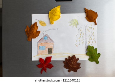 Colorful Felt Autumn Leaves On Refrigerator Door. Children Picture With House And Tree Hanging On Fridge Door. White Kitchen On Background. Diy Fall Autumn Decoration For Home