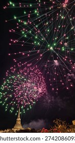 Colorful fireworks exploding in the night sky above a temple pagoda, celebrating a festive event. Bright red and green sparks illuminate the dark sky in a vibrant celebration scene.