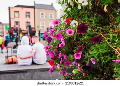 Colorful Flower Bed In Focus In Foreground. Couple Close To Each Other With Travel Suitcase In Old Town Out Of Focus. City Life, Abstract Travel Concept.