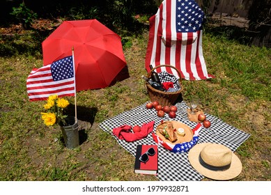 Colorful Fourth Of July Backyard Picnic With Flags, Basket, And Food With A Black And White Checkered Cloth Spread On The Grassy Ground. 