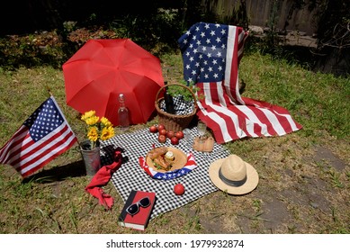 Colorful Fourth Of July Backyard Picnic With Flags, Basket, And Food With A Black And White Checkered Cloth Spread On The Grassy Ground. 