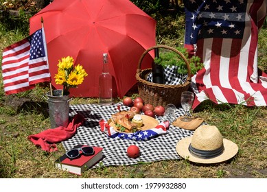 Colorful Fourth Of July Backyard Picnic With Flags, Basket, And Food With A Black And White Checkered Cloth Spread On The Grassy Ground. 