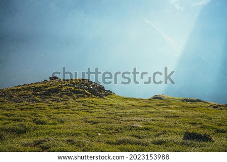 Colorful green landscape with rocks and hills on background of giant mountain wall in sunlight. Minimalist vivid sunny scenery with sun beams and solar flare. Minimal alpine view. Scenic minimalism.