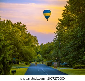 Colorful Hot Air Balloon Flying Over Midwestern Suburban Street At Sunset In Summer; Small Figure Of Walking Woman Underneath