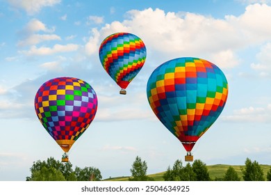 Colorful hot air balloons flying over the green hills and trees at sunset. Rainbow colored balloons in the blue sky. Beautiful summer landscape. 