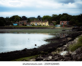 Colorful houses line the coastline, reflecting in calm waters. The rocky beach and gentle waves create a tranquil atmosphere under a cloudy sky. - Powered by Shutterstock