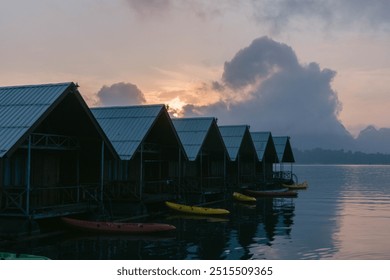 Colorful kayaks rest quietly on the calm water as wooden cabins reflect the soft hues of a tranquil sunrise over the lake, creating a peaceful atmosphere. - Powered by Shutterstock