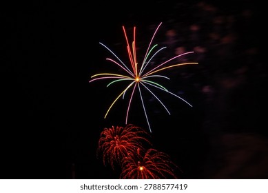 Colorful long exposure fireworks exploding in the night sky with vibrant rainbow light trails and red sparkling bursts against a dark background.