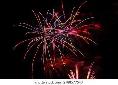 Colorful long exposure fireworks exploding in the night sky with vibrant rainbow light trails and red sparkling bursts against a dark background.