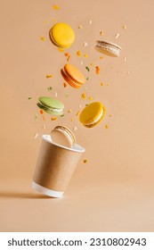 Colorful macarons with crumbs flying out of the paper coffee cup on the  beige backdrop. Levitation.