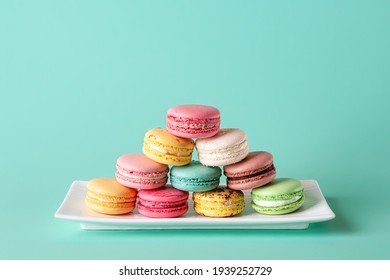 Colorful macaroons pyramid on white rectangular plate against pastel turquoise background. Mockup of greeting card with sweet multicolored macaron cakes. Delicious almond meringue cookies. Copy space.