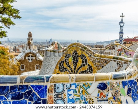 Colorful mosaics on fence of serpentine bench in main square below the Hypostyle of Parc Guell, urban cityscape against cloudy sky in background, sunny day in Barcelona, Catalonia, Spain