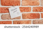 A colorful note saying "Thank you" with a smiley face is pinned to a rustic brick wall, conveying a cheerful message of gratitude and positivity in an urban setting.