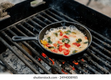 A colorful omelette cooked in a cast iron skillet over a campfire grill. The omelette is filled with fresh vegetables, including bell peppers, tomatoes, and spinach, creating a vibrant and appetizing 